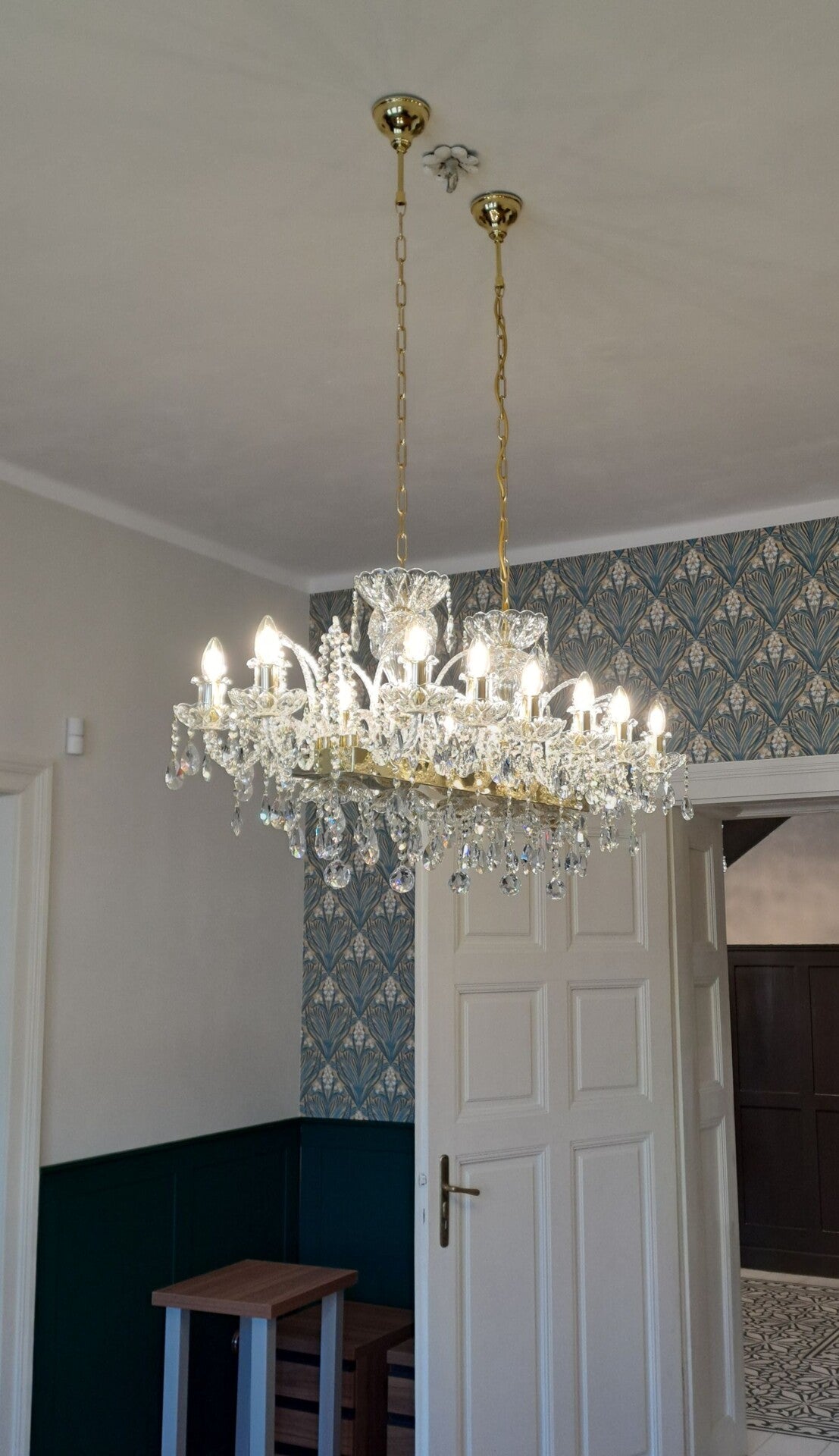 Lustre suspendu par deux chaînes dorées devant un mur à papier peint floral bleu.