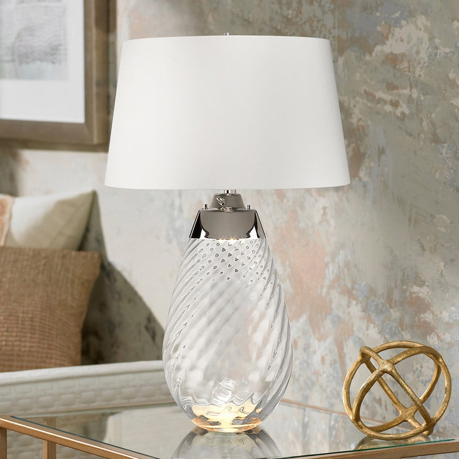Lampe allumée posée sur une table en verre, coussins et tableau en arrière-plan
