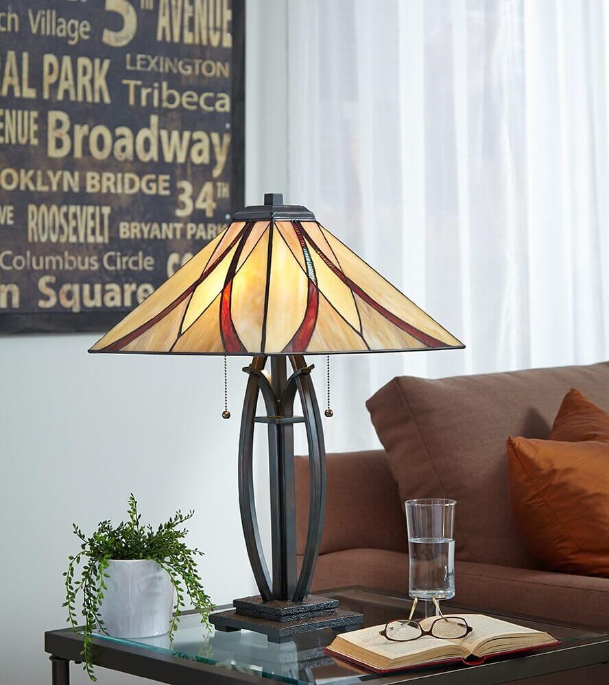 Lampe allumée sur table basse devant un canapé, avec plante, livres et verre d’eau.