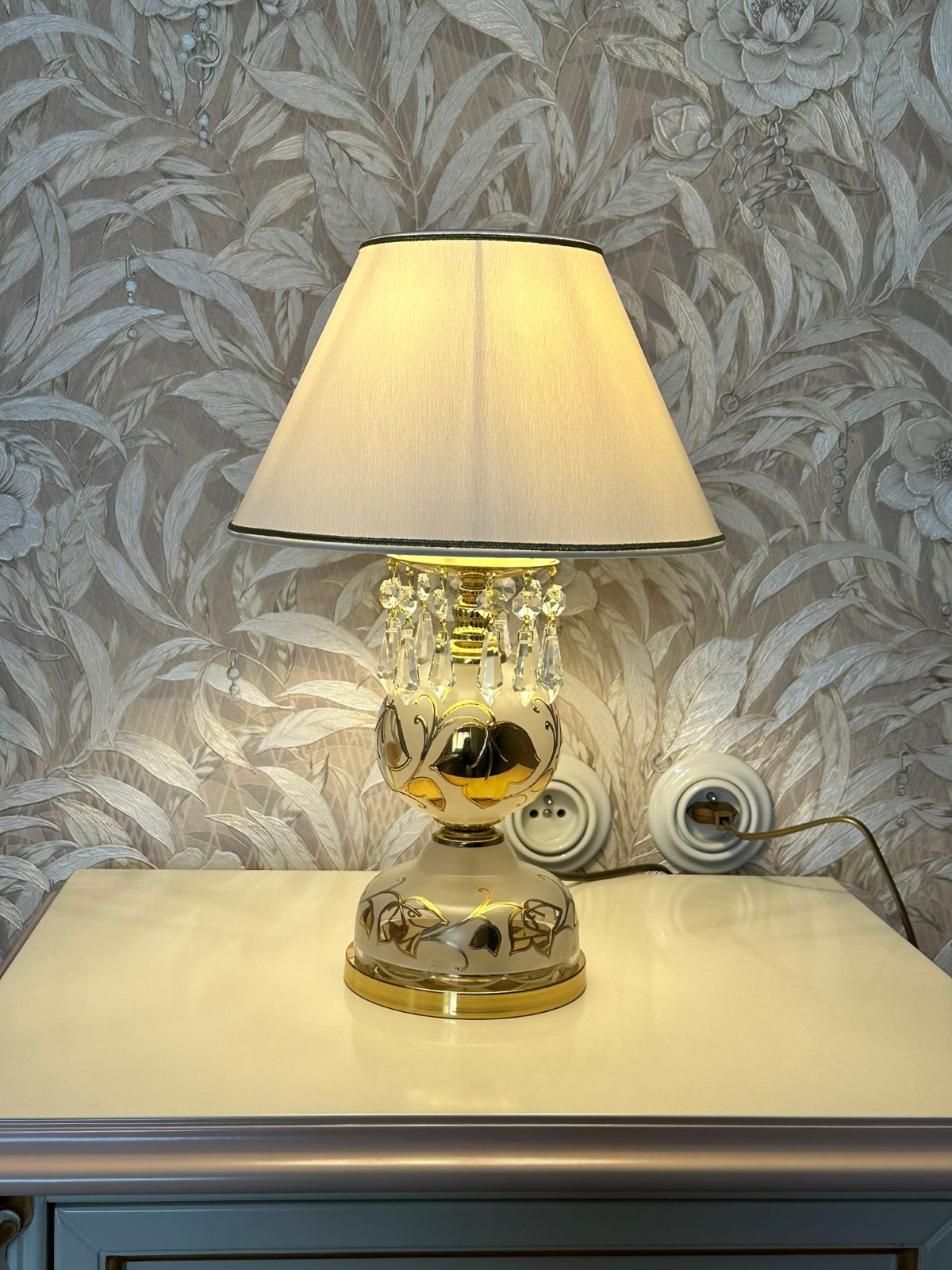 Mise en ambiance de la lampe allumée posée sur une commode devant papier peint floral