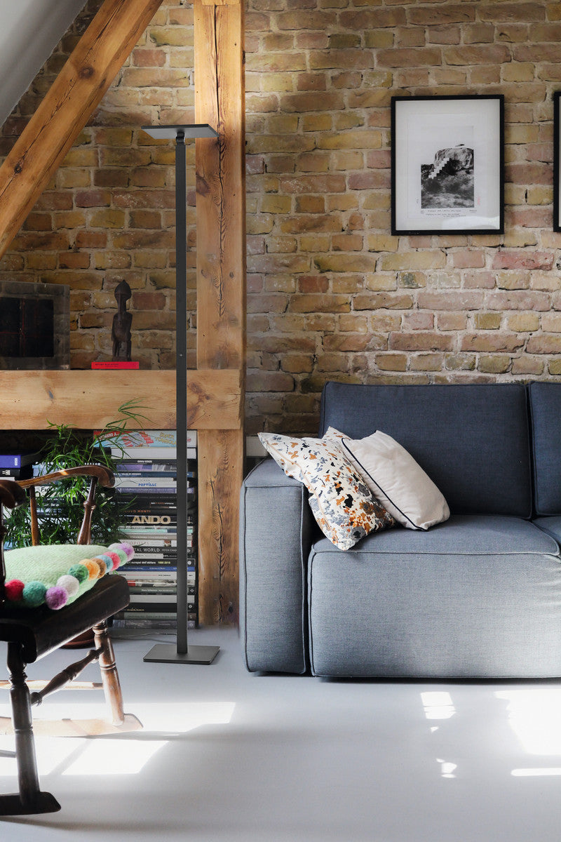 Dans un salon, lampadaire noir dressé près d'un canapé gris et d'un mur de briques.