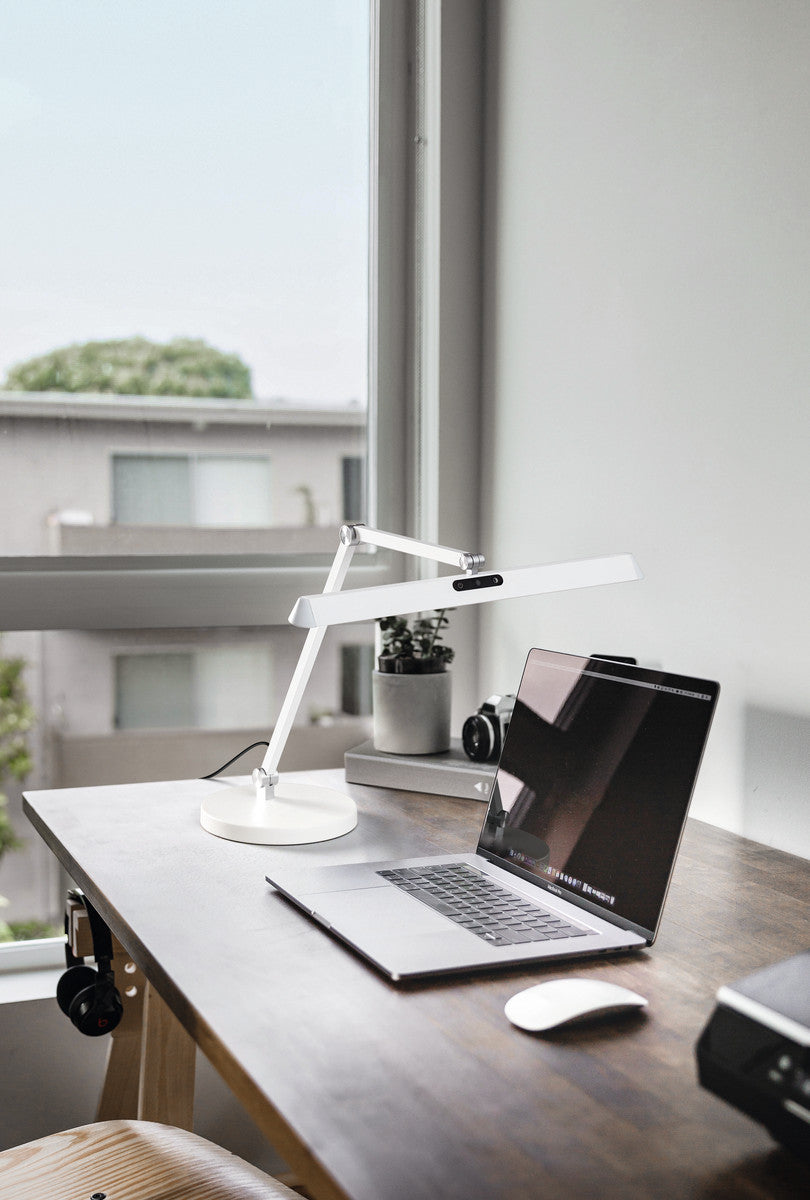 Lampe blanche allumée posée sur un bureau en bois, ordinateur et fenêtre en arrière-plan
