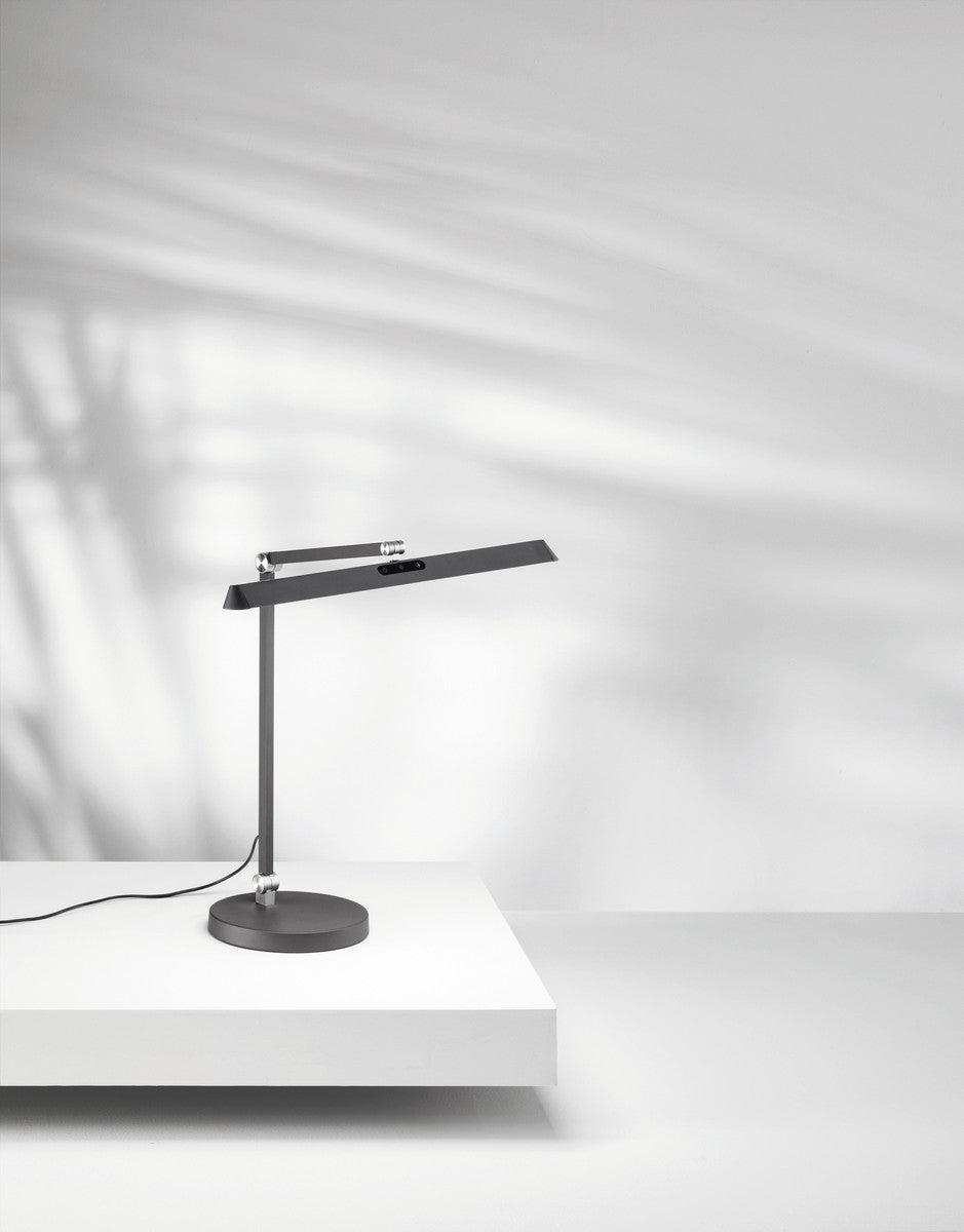 Lampe de bureau anthracite sur surface blanche, ombres portées et câble visible.
