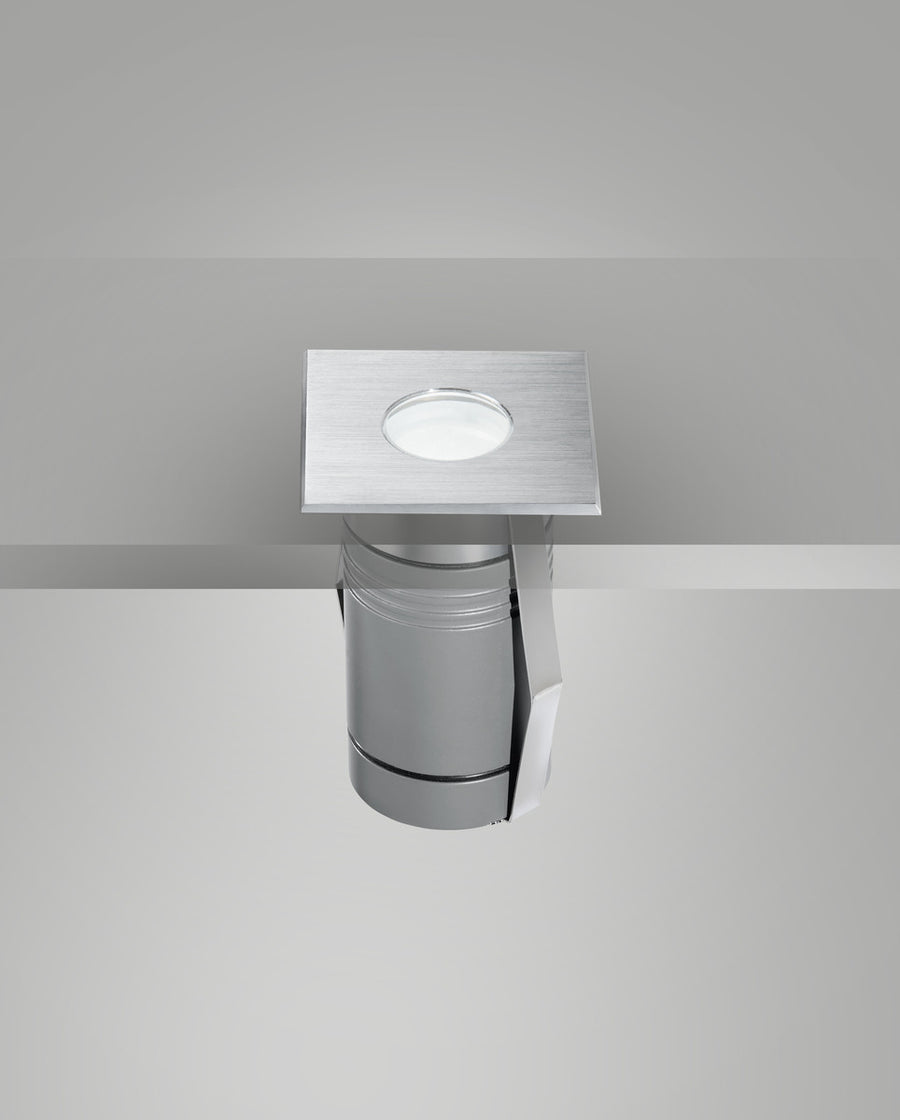 Vue montrant le luminaire encastré dans une surface grise, platine affleurante et lentille centrale.