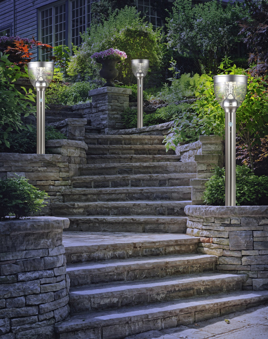 Trois luminaires installés le long d’escaliers en pierre, jardin paysager au crépuscule.
