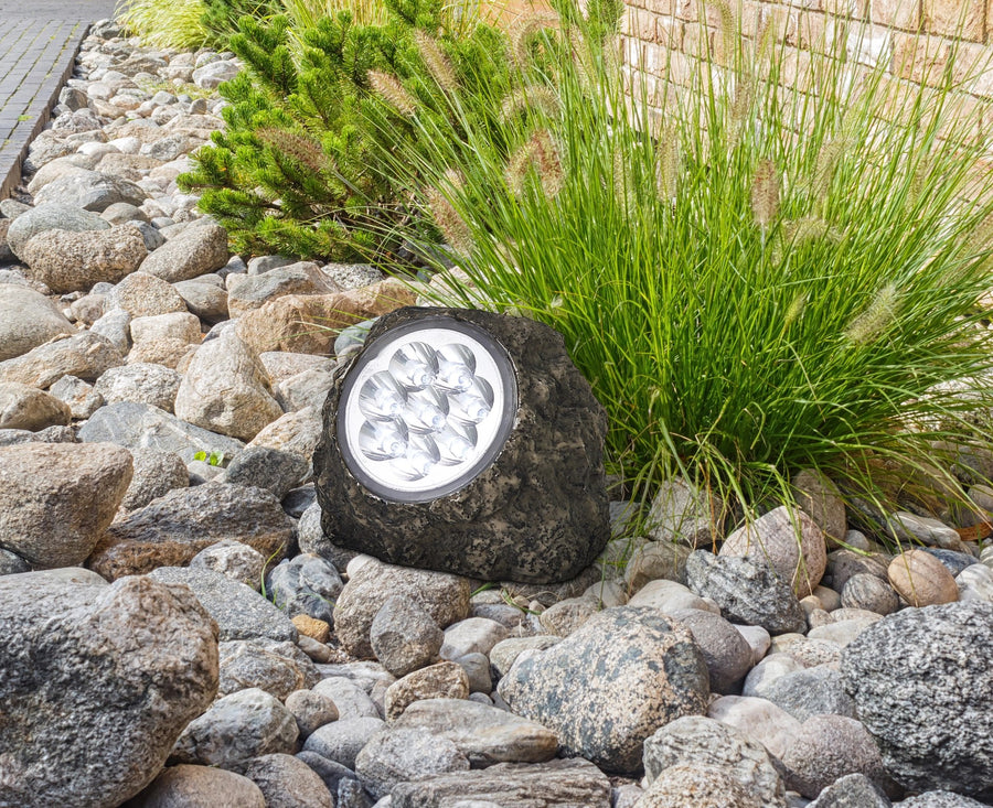 Luminaire solaire roche grise posé sur graviers, entouré de pierres et d'herbes.