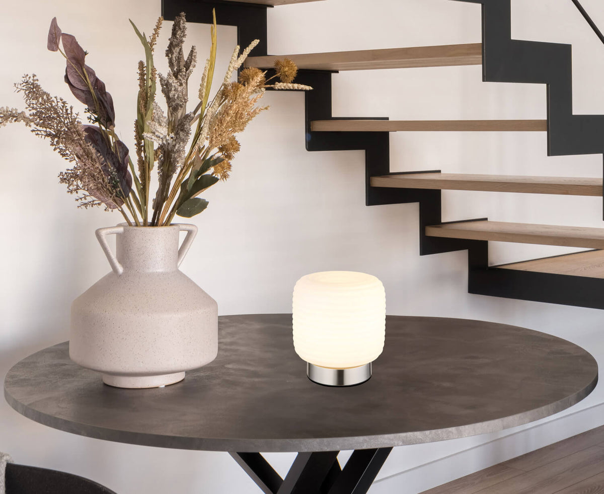 Lampe à poser éclairée sur table ronde en intérieur, près d'un vase et d'un escalier.