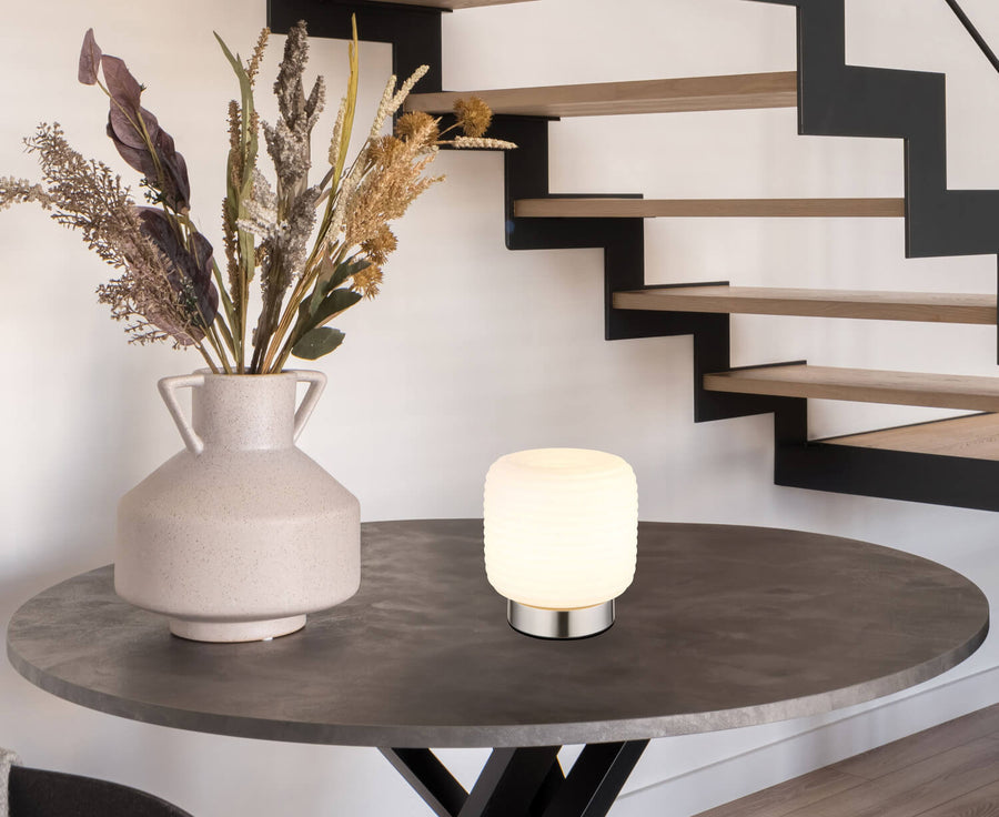 Lampe à poser éclairée sur table ronde en intérieur, près d'un vase et d'un escalier.