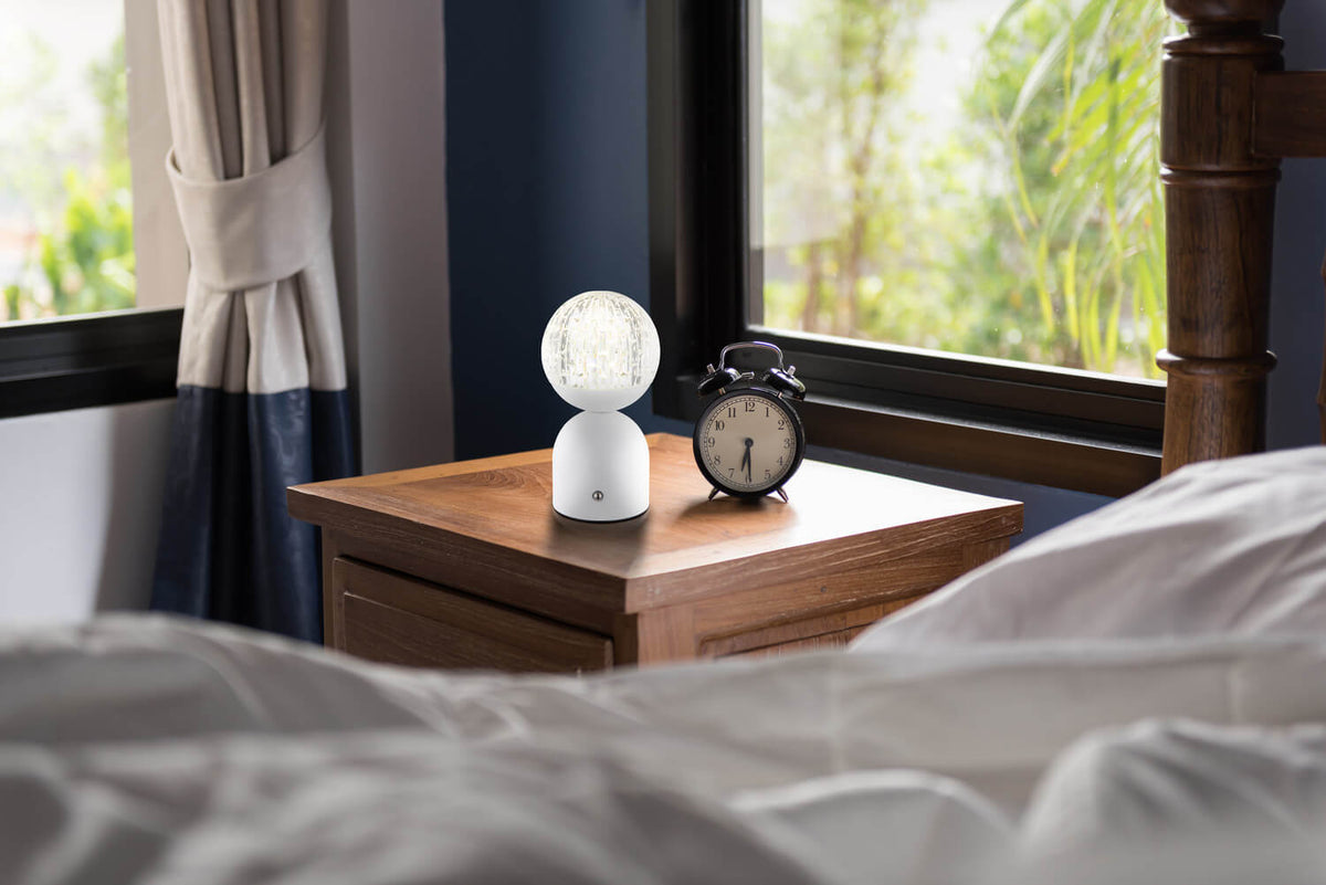 Lampe à poser blanche allumée sur une table de nuit en bois près du lit.