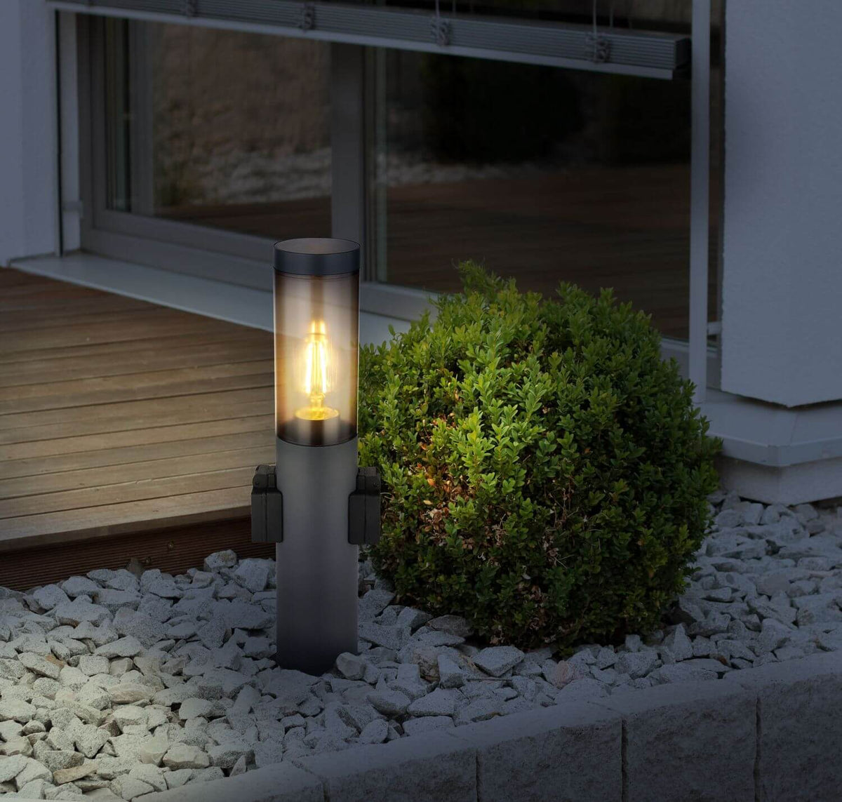 Mise en ambiance nocturne du luminaire près d'une terrasse en bois et d'un arbuste.