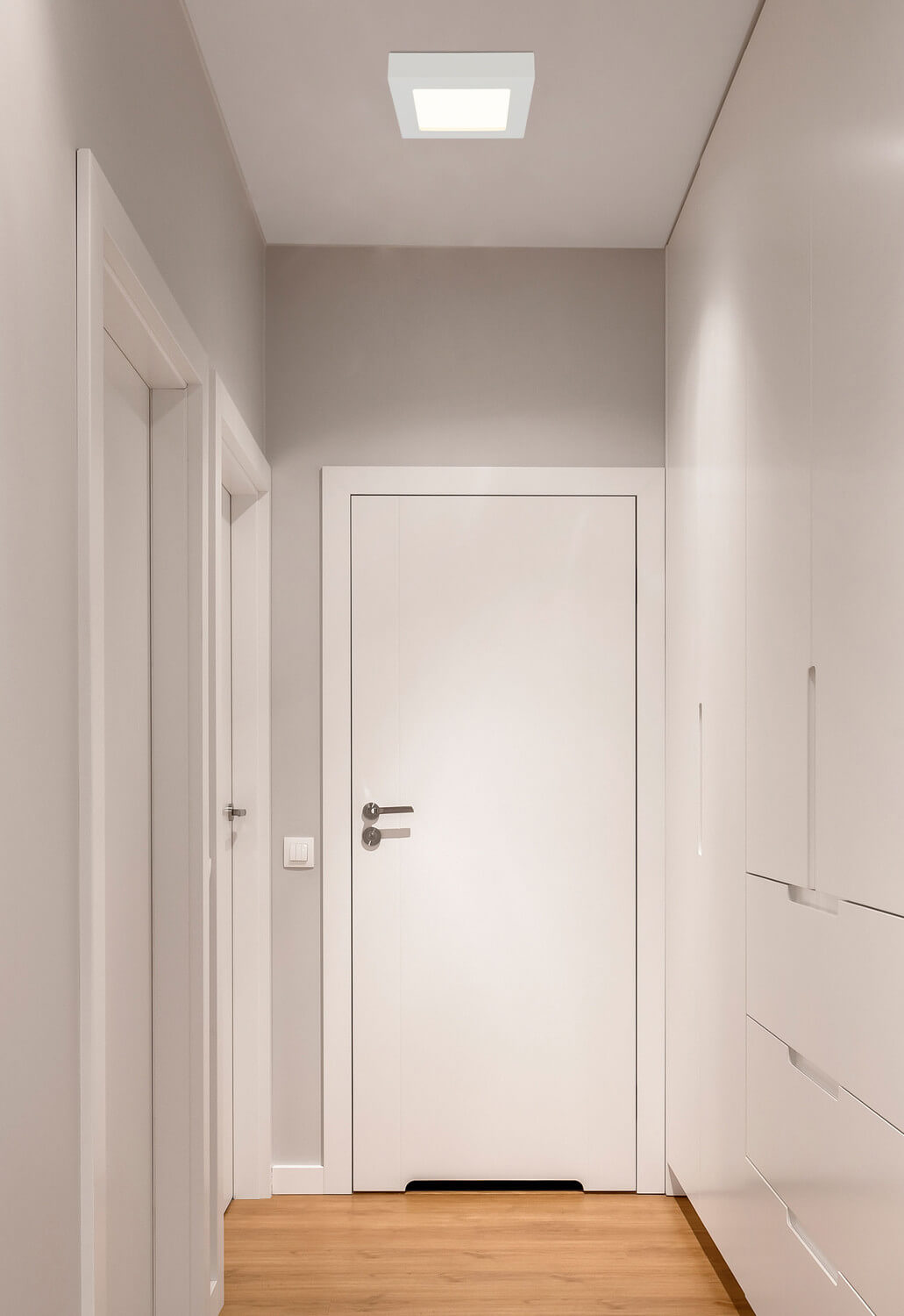 Mise en ambiance dans un couloir blanc, le plafonnier carré éclaire le passage.