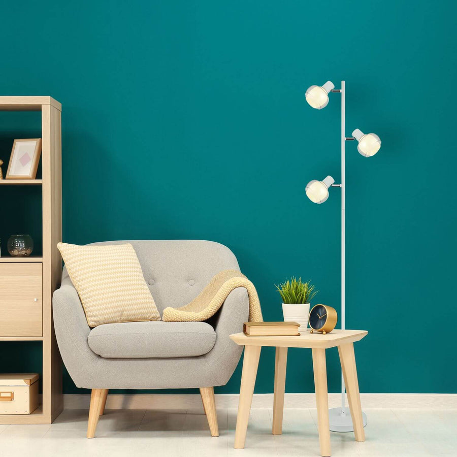 Lampadaire blanc mat installé dans un salon au mur turquoise, près d'un fauteuil gris.