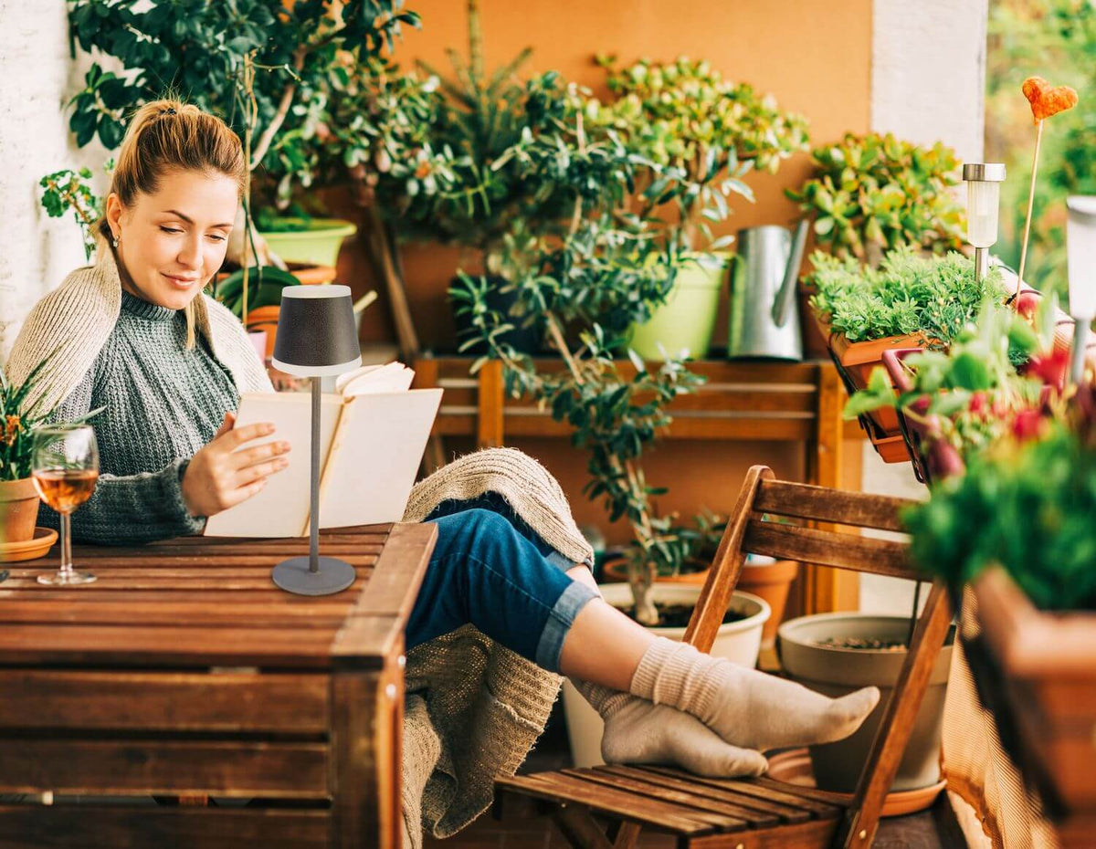 Mise en ambiance : lampe posée sur une table en bois, femme lisant sur une terrasse fleurie.