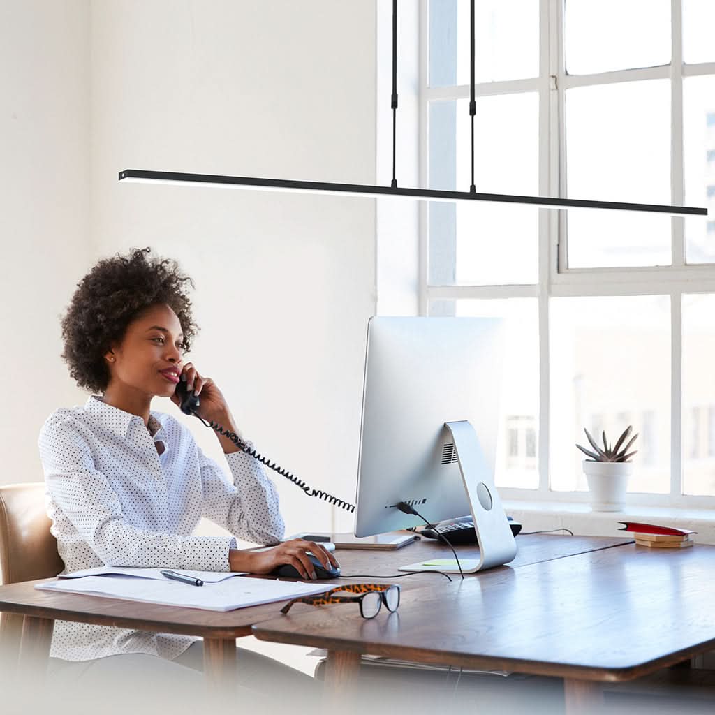 Scène de bureau : femme téléphonant devant ordinateur, luminaire linéaire noir suspendu.