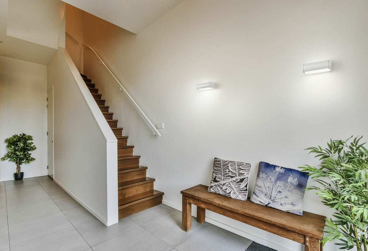 Mise en ambiance dans une entrée avec deux appliques murales allumées, escalier et banc en bois.