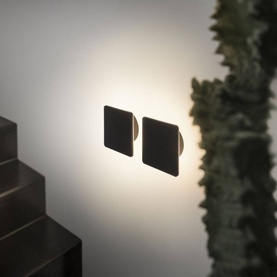 Deux appliques carrées noires vues de profil, halo lumineux diffus sur le mur
