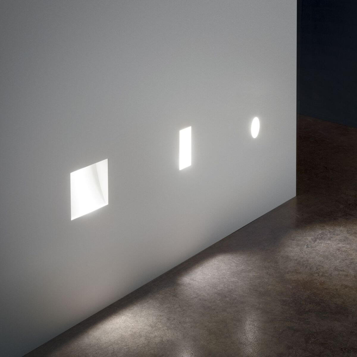 Mise en ambiance de trois spots encastrés allumés sur un mur blanc, lumière rasante