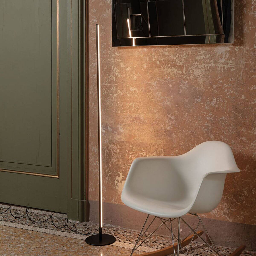 Lampadaire vertical allumé diffusant une lumière chaude sur un mur texturé et une chaise blanche.
