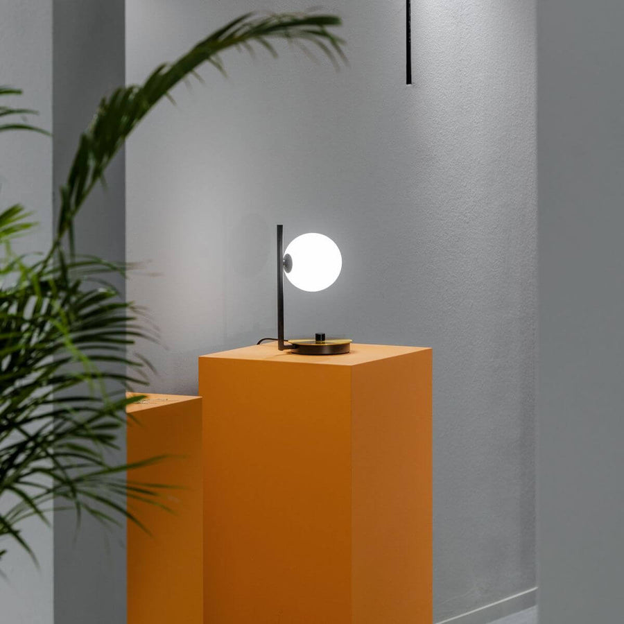 Lampe à poser allumée sur un piédestal orange, feuillage de palmier flou au premier plan.