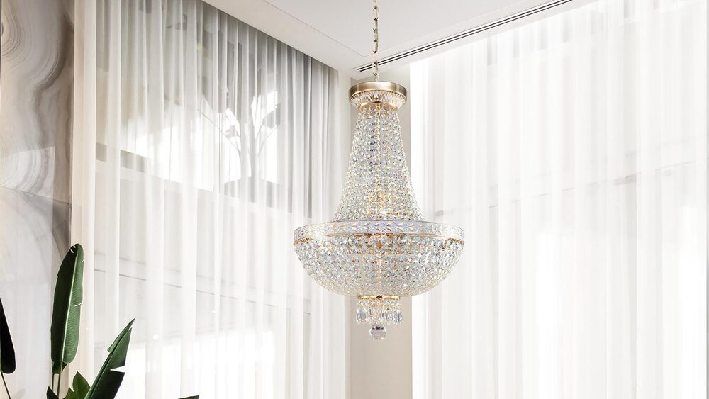 Lustre en cristal doré suspendu devant de grands rideaux clairs, dans un intérieur lumineux et aéré.