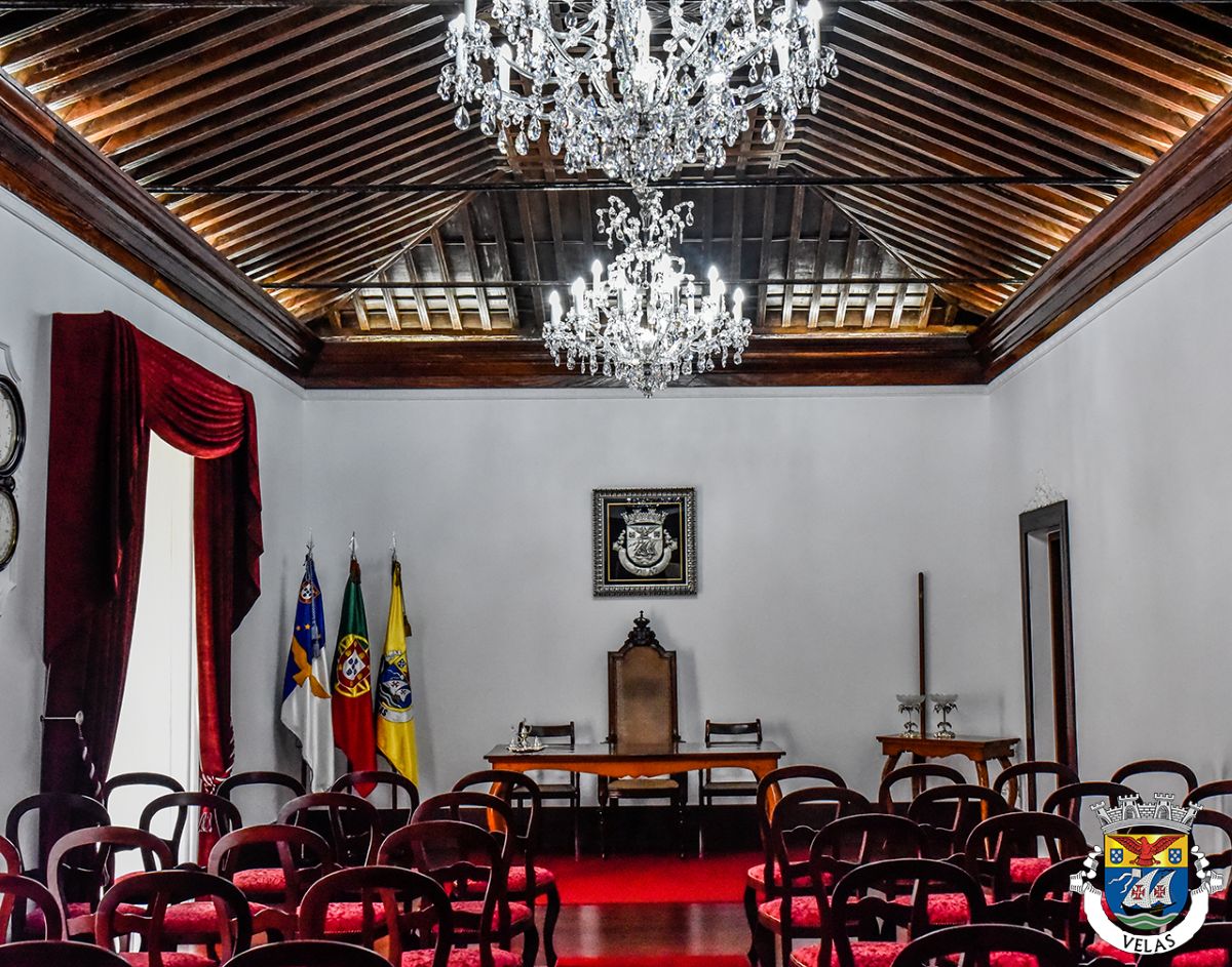 Salle officielle au plafond de bois où le lustre éclaire les fauteuils rouges et les drapeaux
