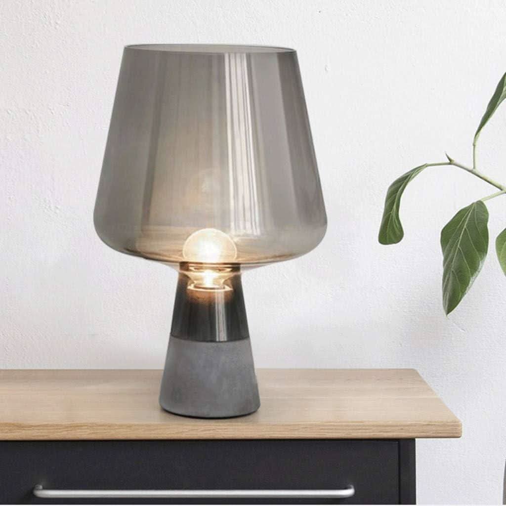 Lampe à poser allumée sur une commode en bois, mur blanc et feuillage à droite.