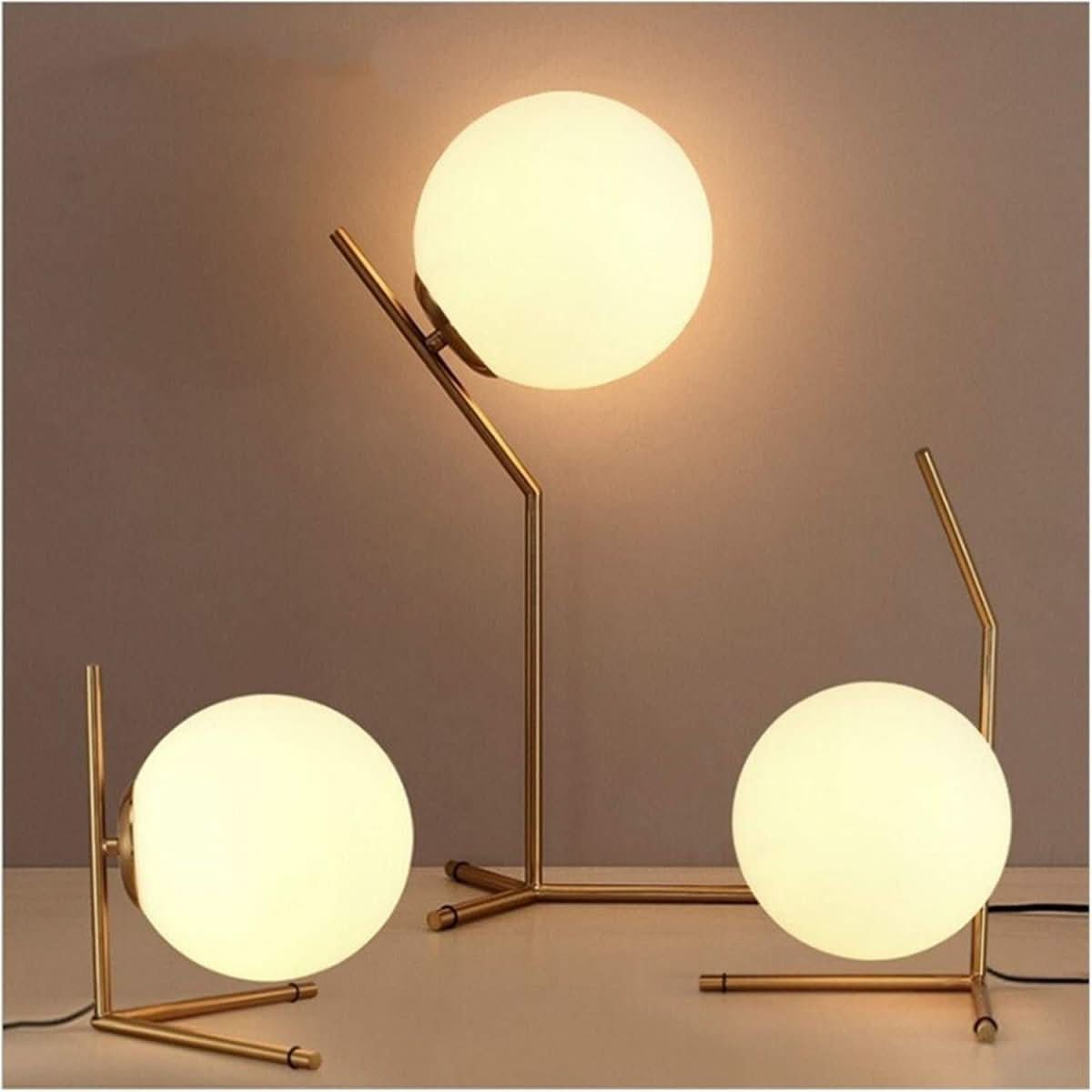 Trois lampes boule dorées posées, pieds trépied fins, sphères opalines allumées sur fond beige.