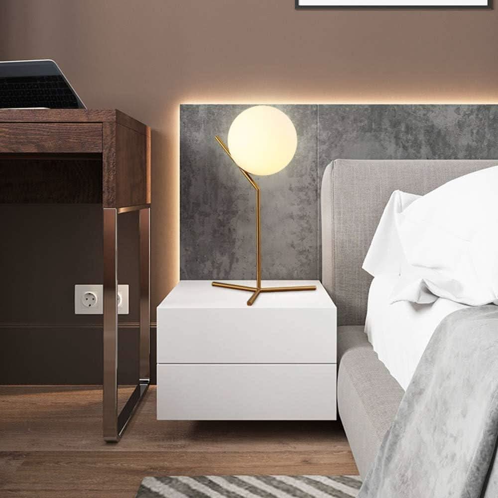 Lampe boule dorée posée sur une table de nuit blanche, tête de lit grise en arrière-plan.