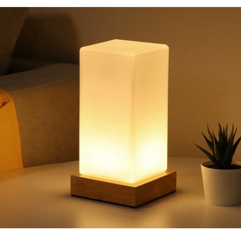 Lampe rectangulaire allumée sur base en bois, posée près d'une plante en pot.