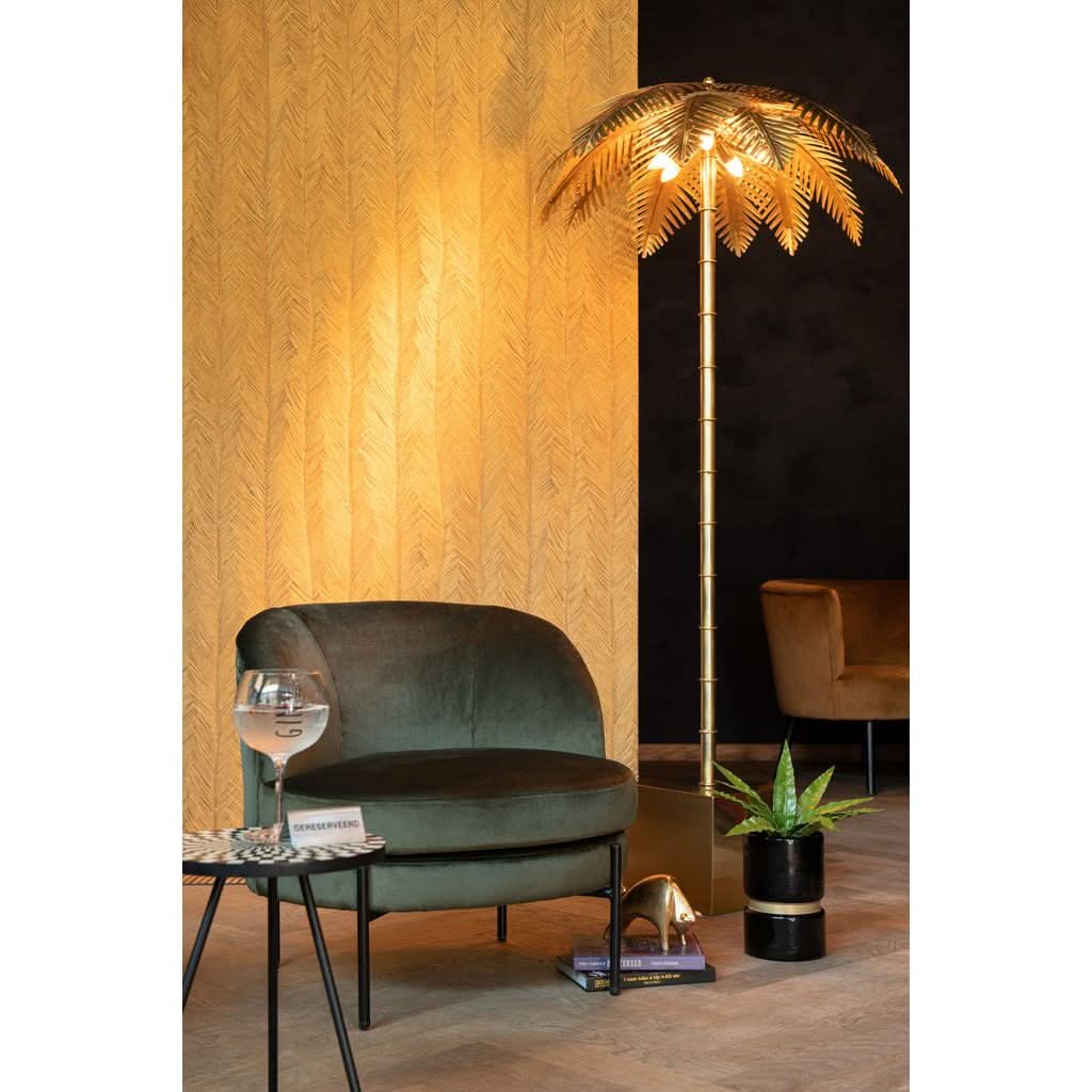 Lampadaire doré allumé devant papier peint doré, fauteuil vert et plante verte.