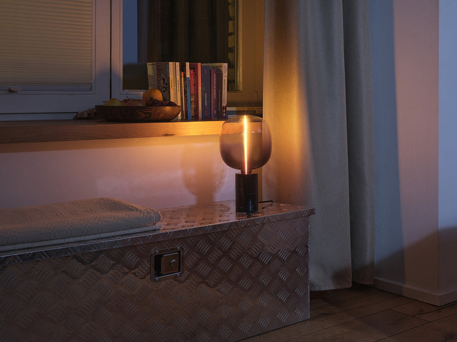 Petite lampe à poser allumée sur coffre métallique, halo chaud près de la fenêtre.