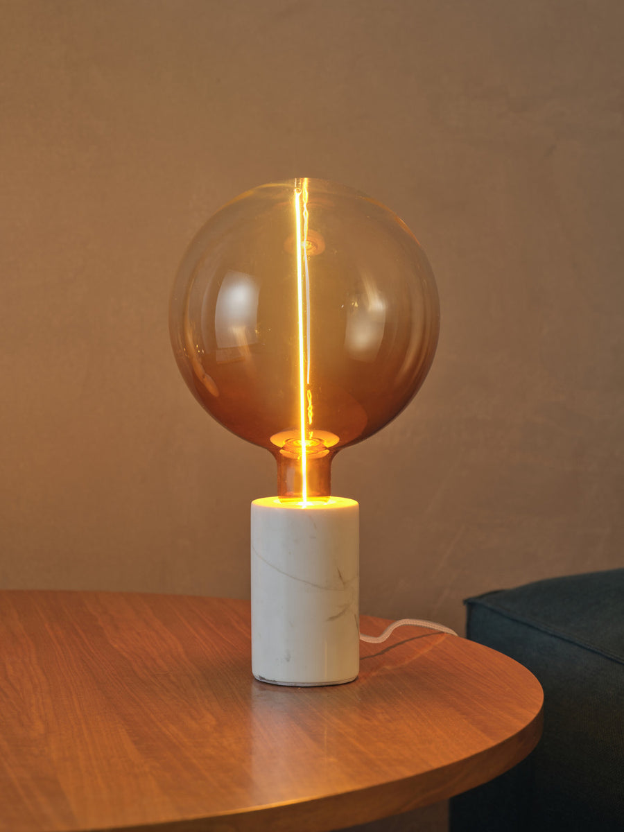Lampe à poser en marbre allumée sur une table en bois, ampoule globe visible.