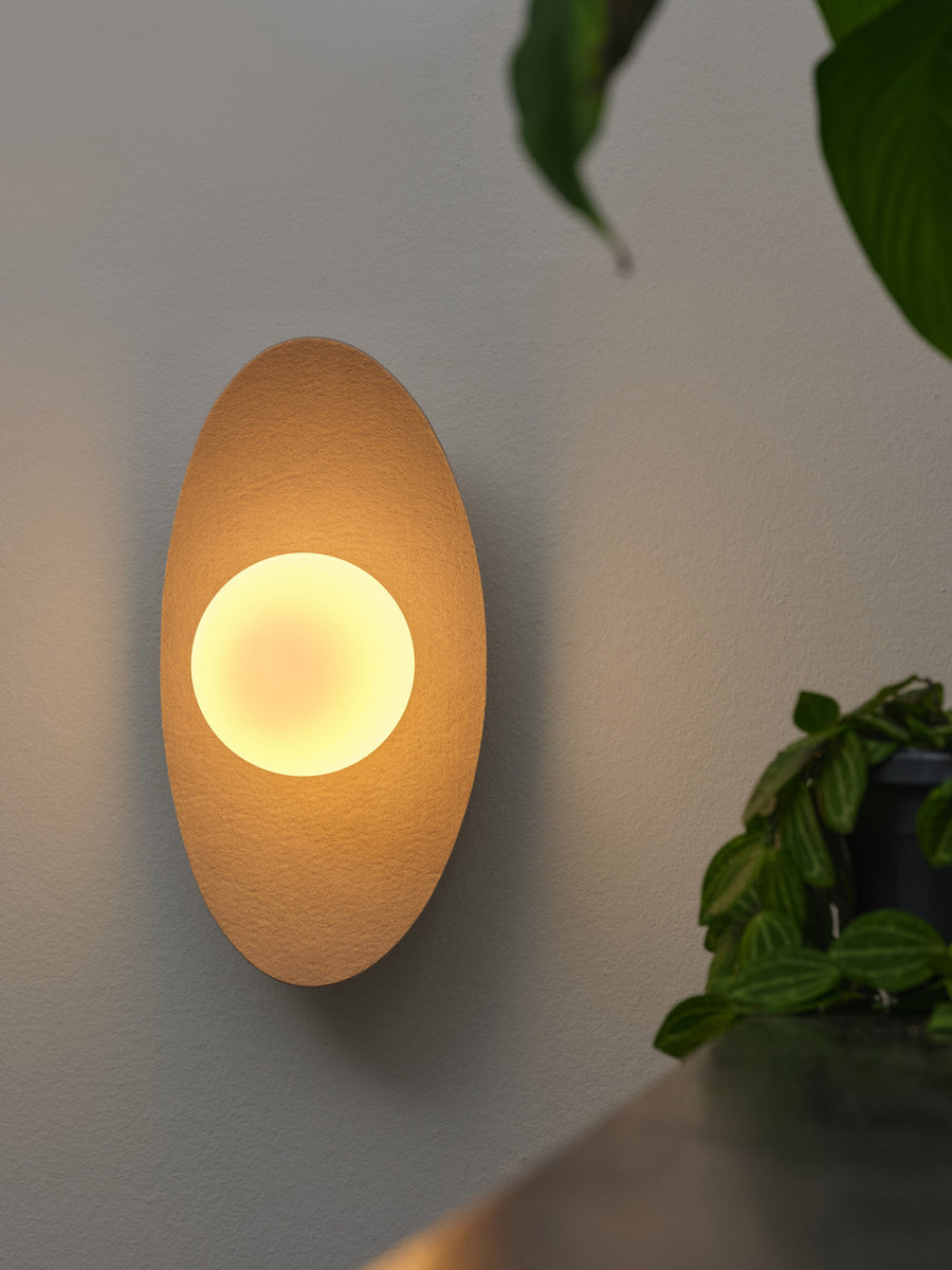 Applique murale beige allumée diffusant une lumière chaude, avec plantes à l’arrière-plan.