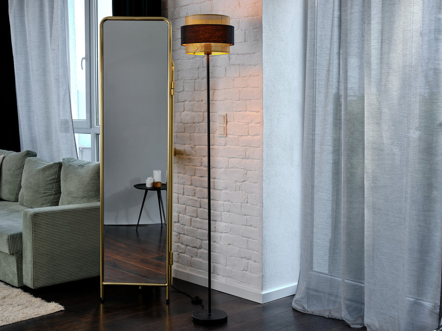 Lampadaire allumé dans un salon, devant un mur de briques blanches, miroir doré et canapé vert.
