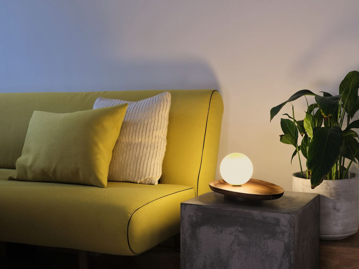 Lampe allumée posée sur table basse en béton, près d’un canapé jaune et d’une plante.