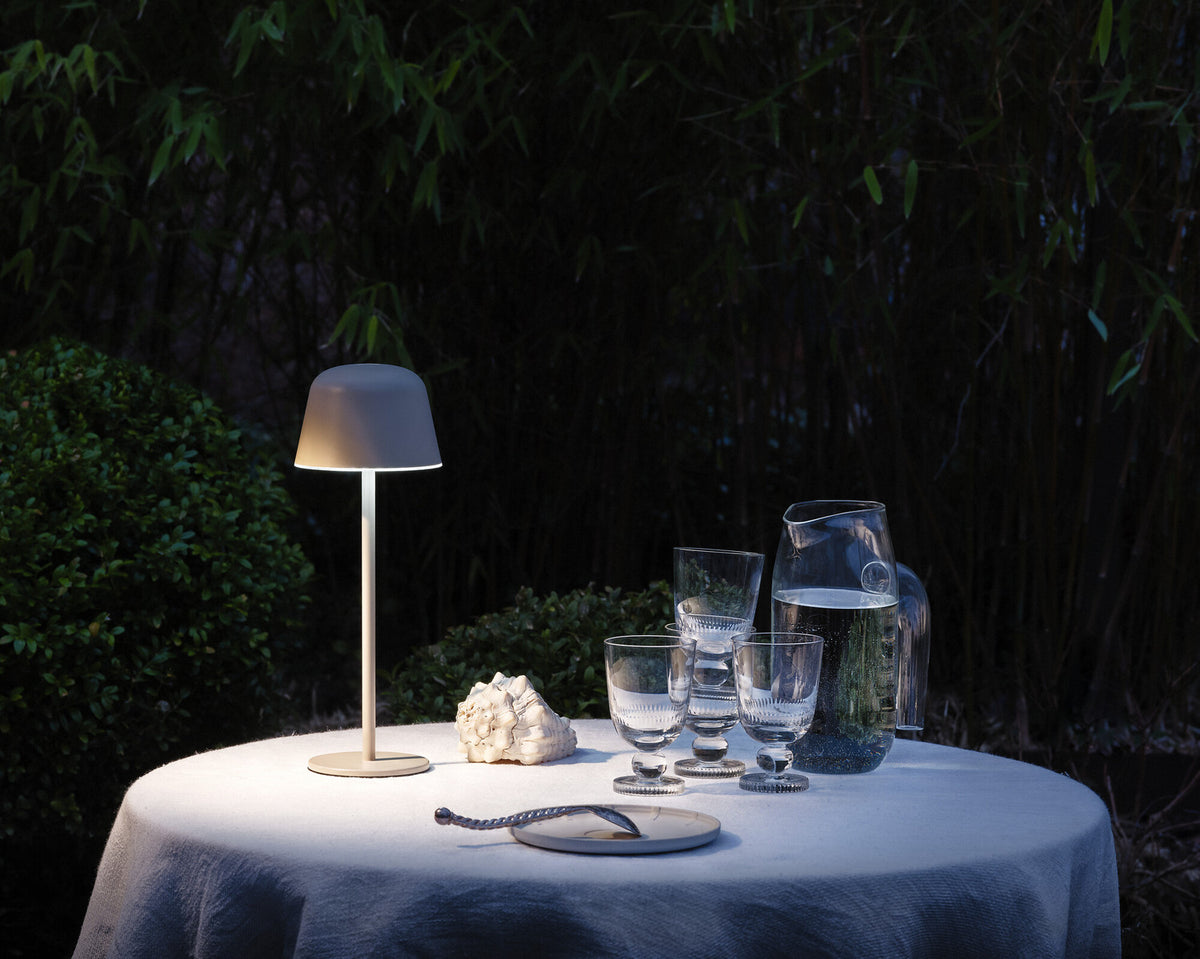 Lampe allumée sur une table ronde dehors, près de verres et d’un pichet.