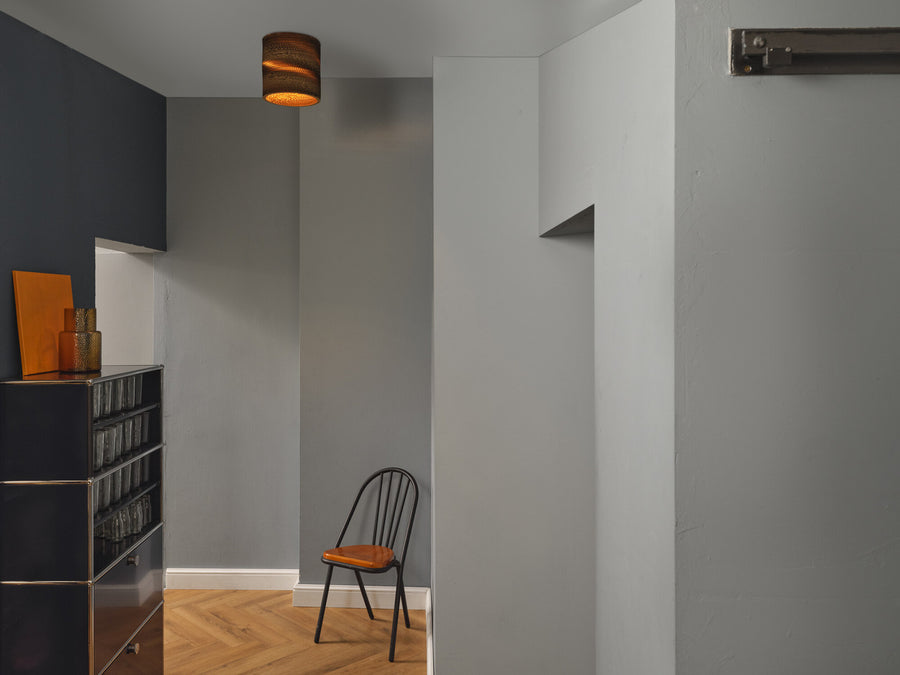 Plafonnier allumé dans un couloir gris, au-dessus d'une chaise noire et d'un meuble.