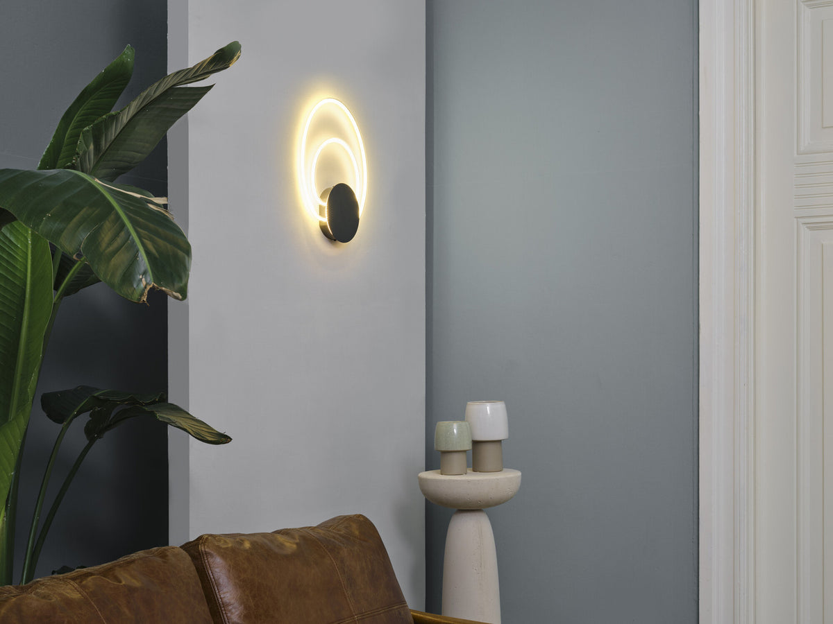 Mise en ambiance dans un salon, applique allumée sur mur gris près d'une plante.