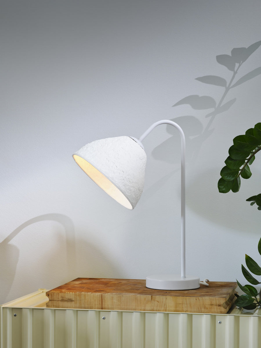 Lampe allumée sur un support en bois, ombre de feuillage sur le mur blanc.