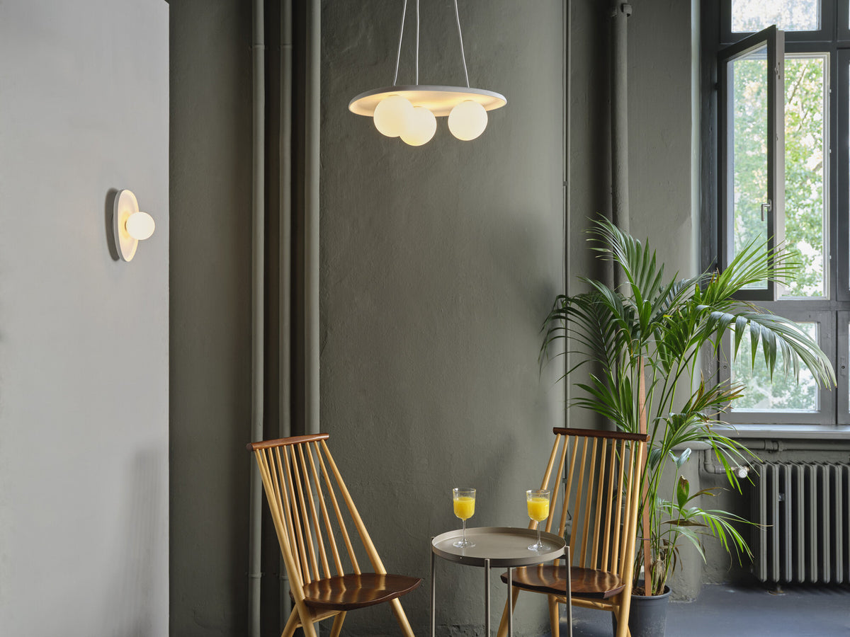 Mise en ambiance intérieure avec l’applique murale allumée, un luminaire suspendu, des chaises et une plante.