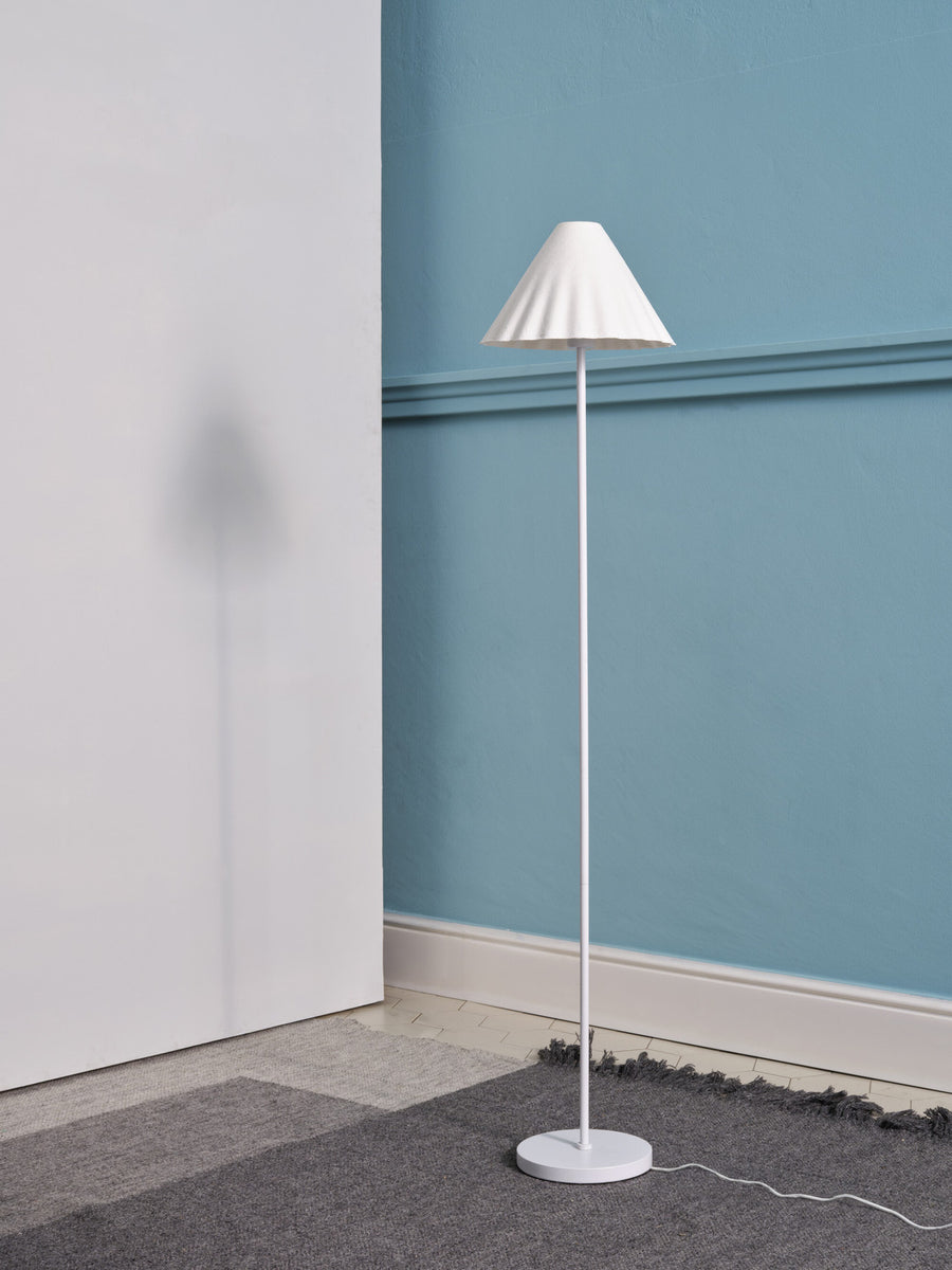 Lampadaire blanc éteint dans un coin de pièce, sur une moquette grise.