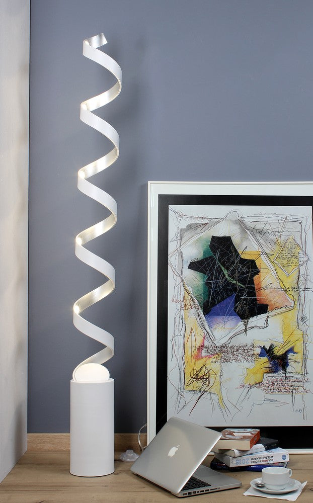 Vue d'ambiance éclairée : lampadaire spiralé allumé, cadre au mur, ordinateur, livres et tasse.
