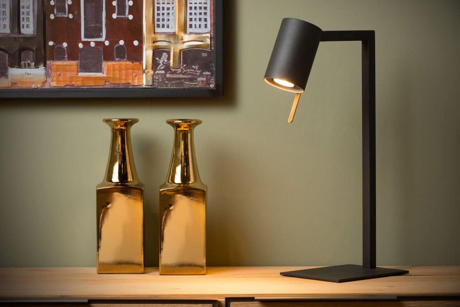 Vue rapprochée de la lampe noire éclairée, posée devant un tableau coloré et des vases dorés.