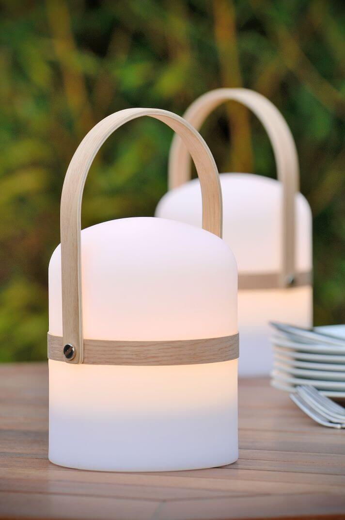 Lampe blanche allumée sur une table en bois, plantes vertes floues et assiettes empilées