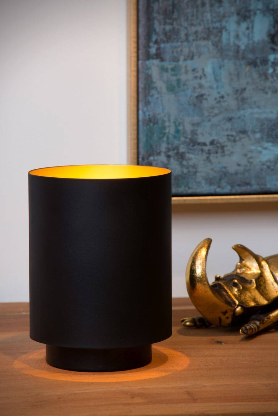 Lampe cylindrique allumée sur une table en bois, éclairant un scarabée doré et un tableau.