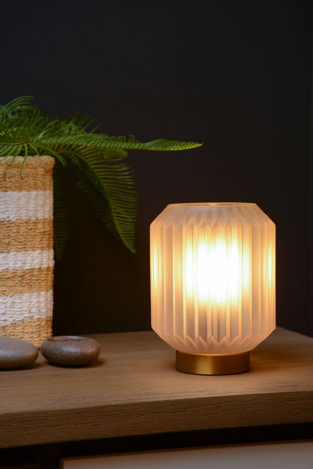 Lampe à poser allumée sur une table en bois, avec plante et pierres décoratives.