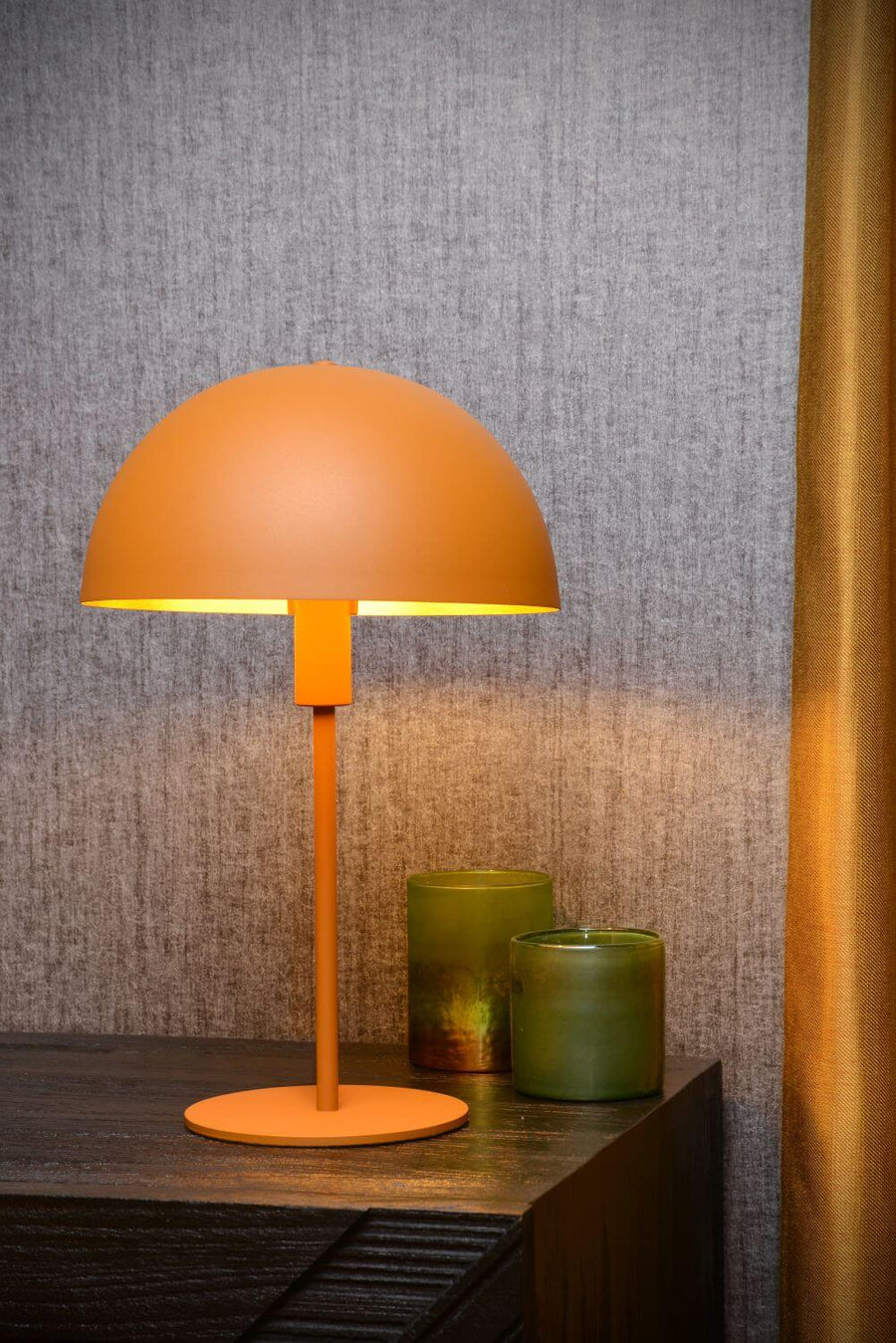 Lampe jaune posée sur commode foncée, deux photophores verts et rideau doré