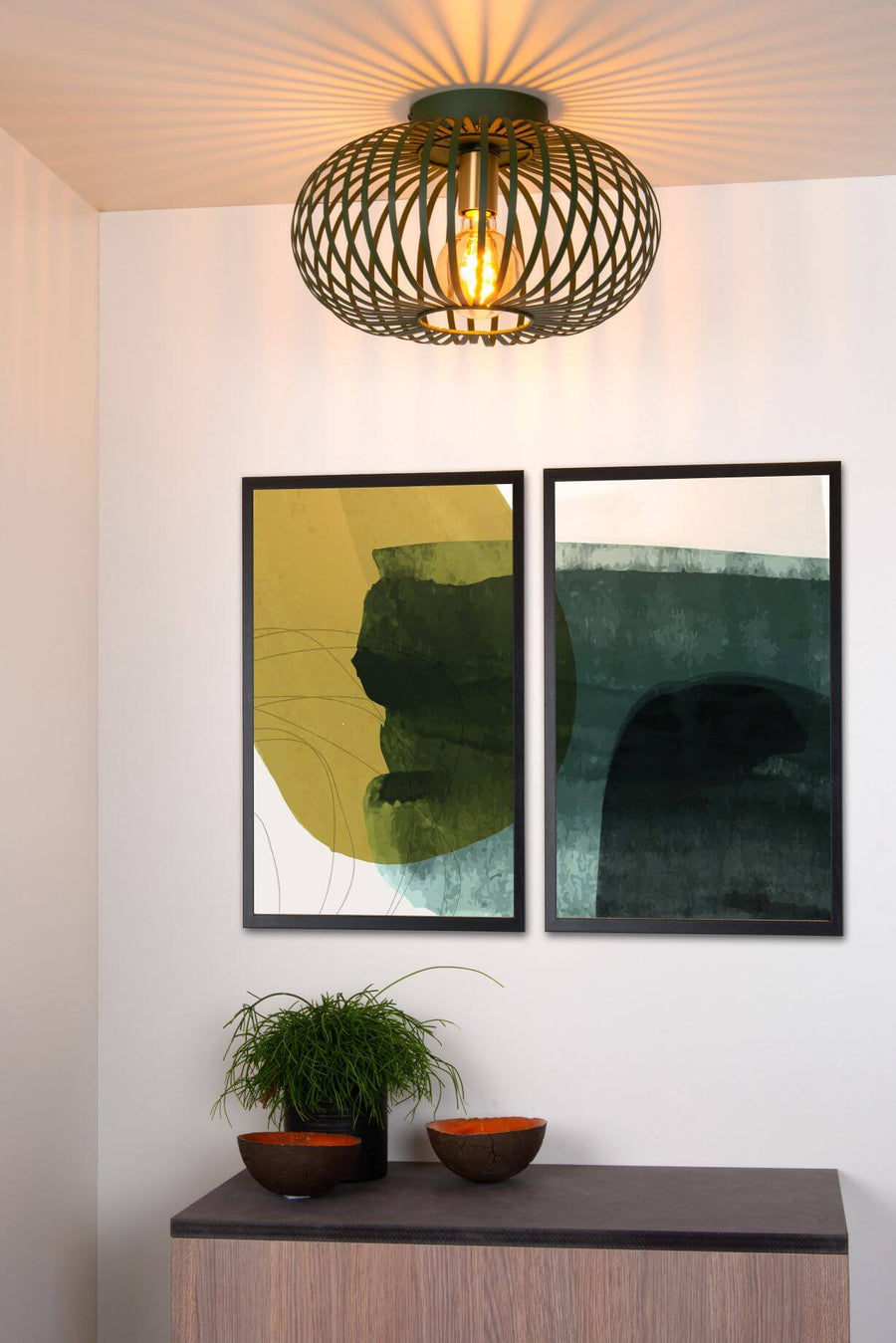 Vue rapprochée du plafonnier vert allumé, avec tableaux et plante en dessous.