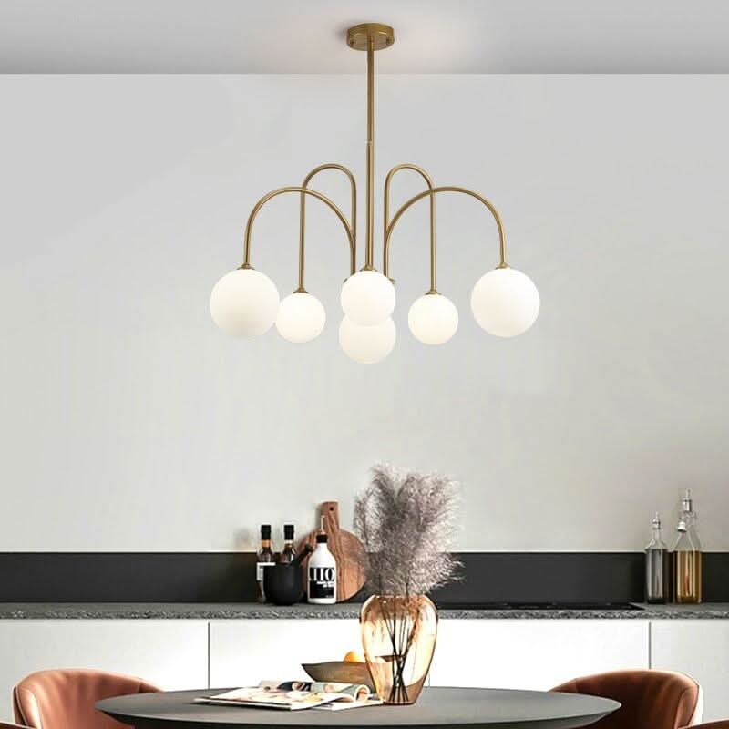 Suspension dorée à globes blancs au-dessus d'une table ronde, vase de fleurs séchées et cuisine.