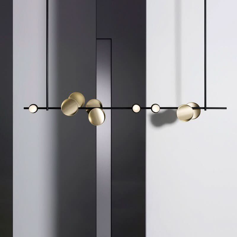 Lustre long suspendu avec barre noire, disques dorés et tiges verticales sur fond géométrique.